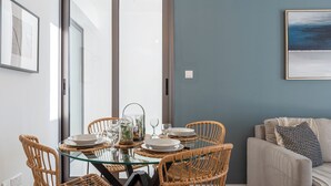 A dining area with a glass table surrounded by wicker chairs, a sofa, and a painting on the wall. - Pauhad 3 - Larnaca Studios & Apts (Larnaca)