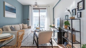 A modern living room with a sofa, a dining table, and chairs, a chandelier, and a balcony with a view. - Pauhad 3 - Larnaca Studios & Apts (Larnaca)
