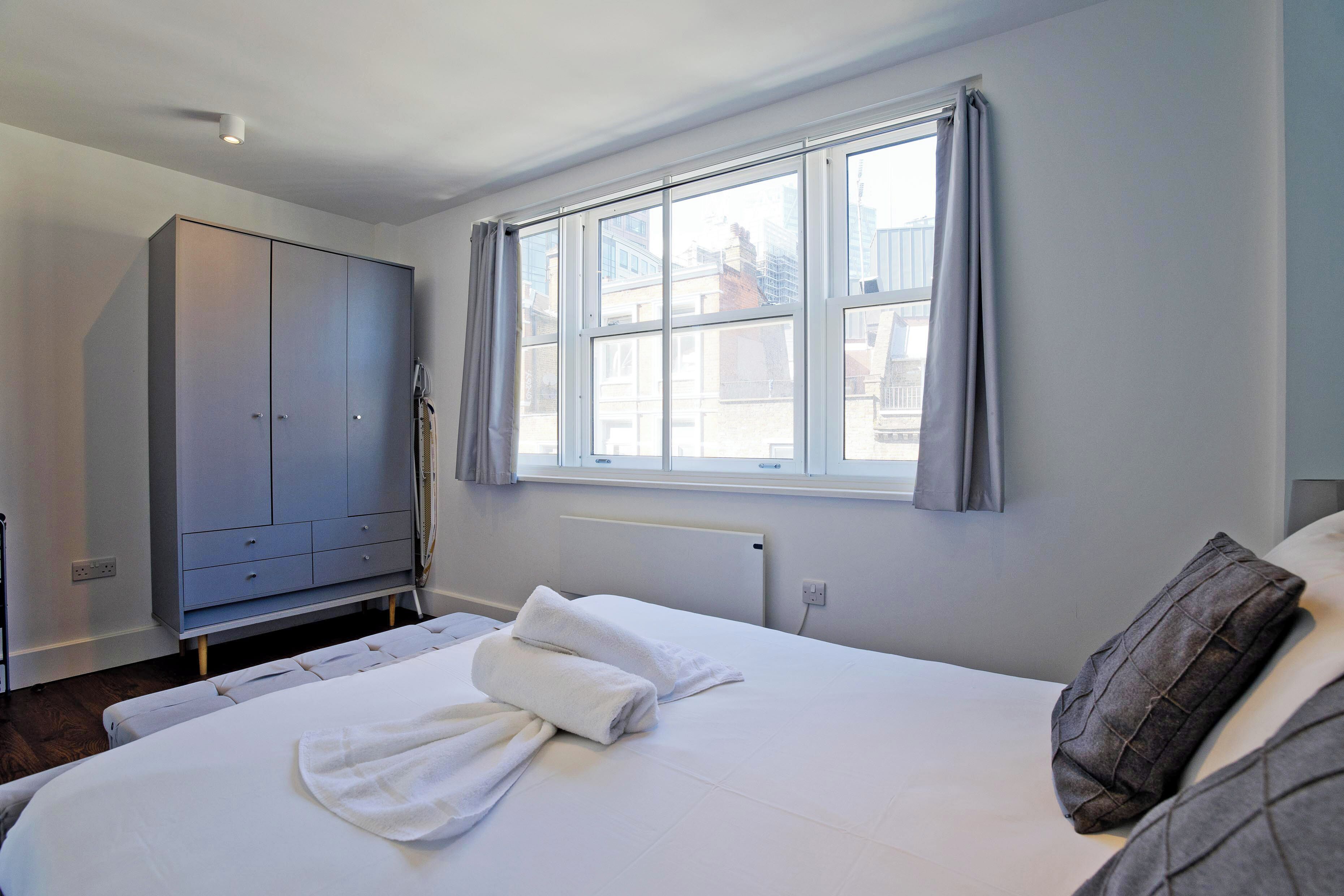 A hotel room with a bed, a wardrobe, a window with curtains, and a view of buildings outside.