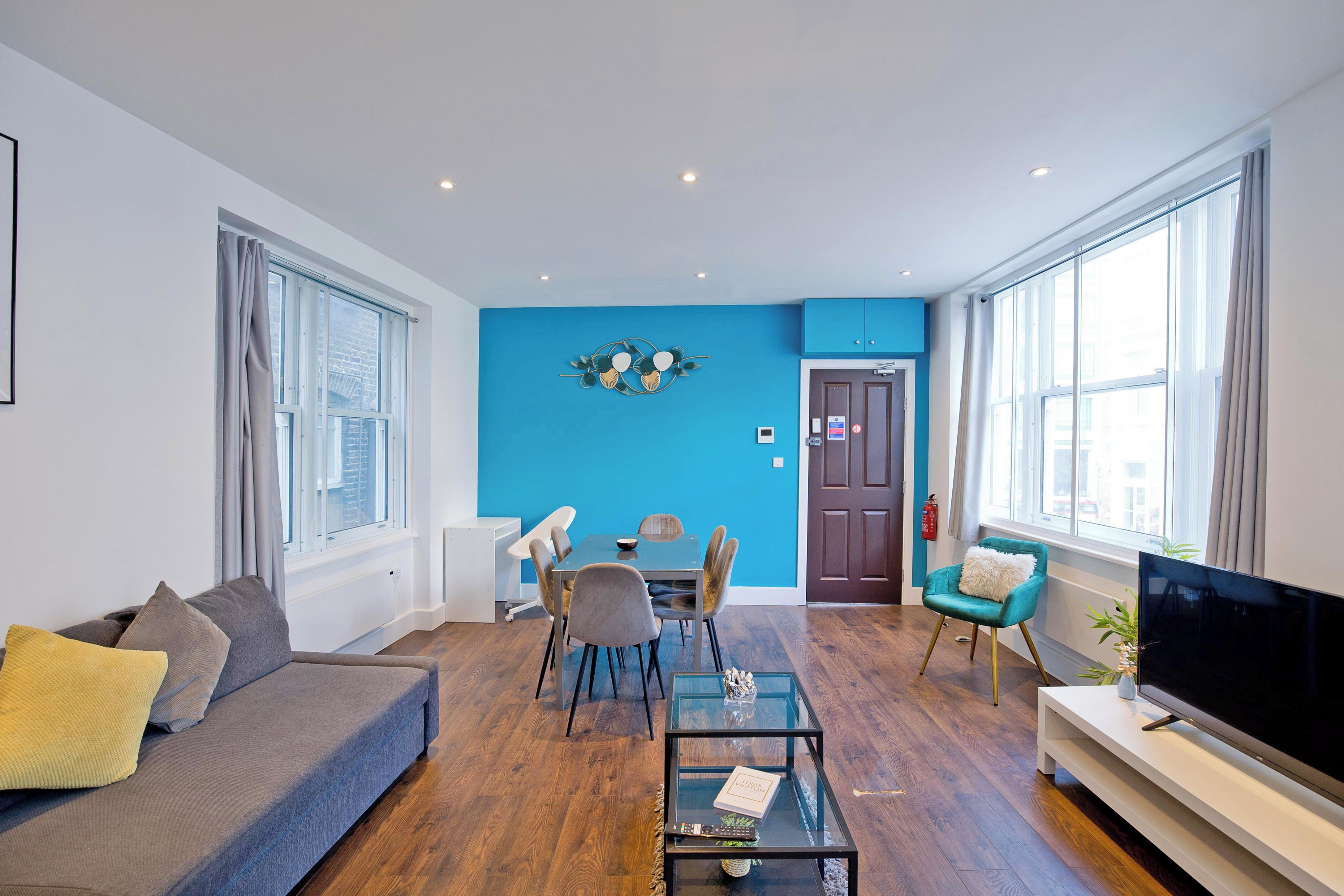 Una sala de estar moderna con una pared azul de acento, un sofá, una mesa de comedor con sillas, un televisor sobre un mueble bajo y ventanas amplias.