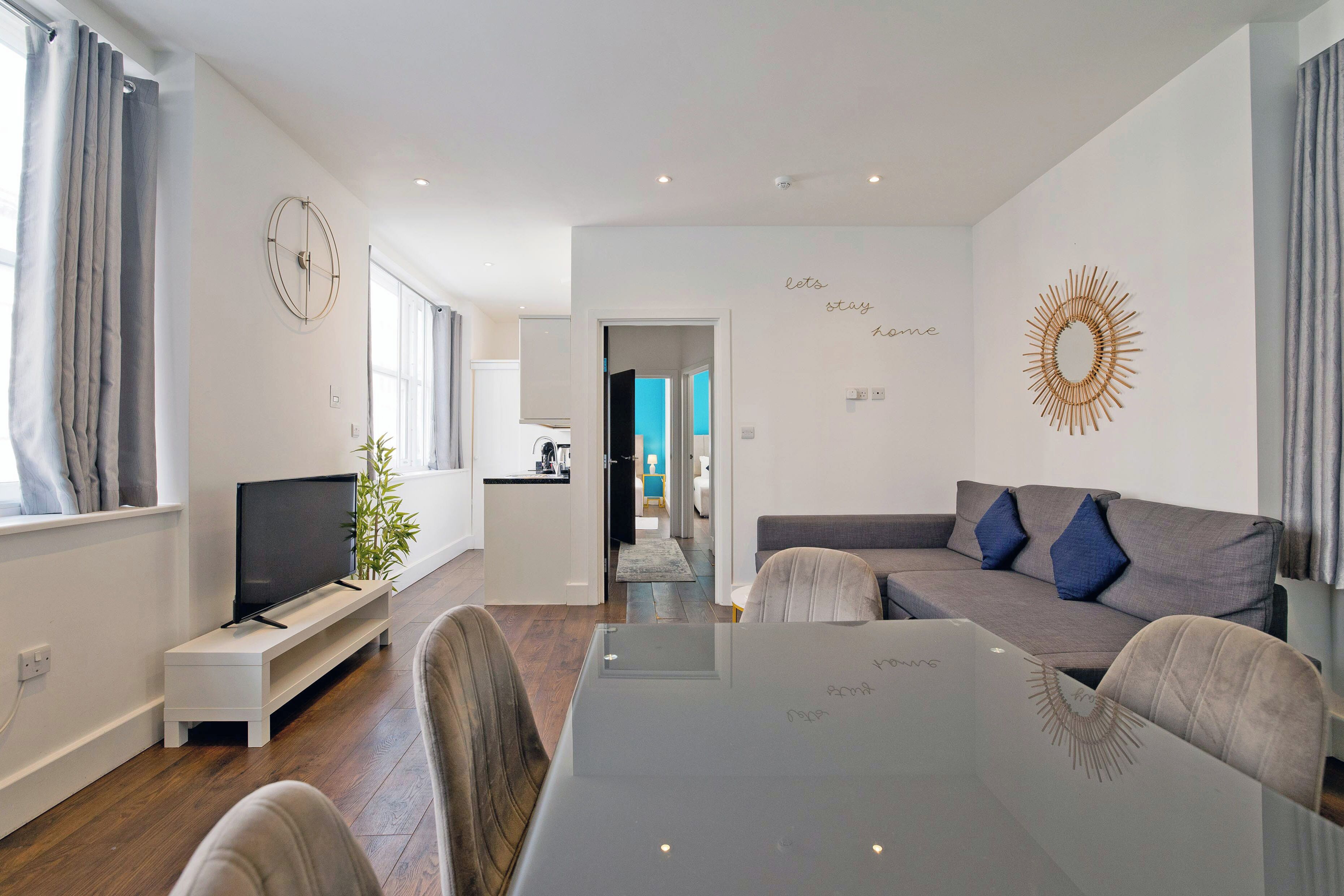 Ein modernes Wohnzimmer mit einem Flachbildfernseher, einem grauen Sofa mit blauen Kissen, einem Glastisch und einem dekorativen Wandbild im Sonnenstrahl-Design.