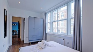 A hotel room with a large bed, a wardrobe, a window with curtains, and a view of buildings outside. - Imperial Middlesex Street Apartments (London)