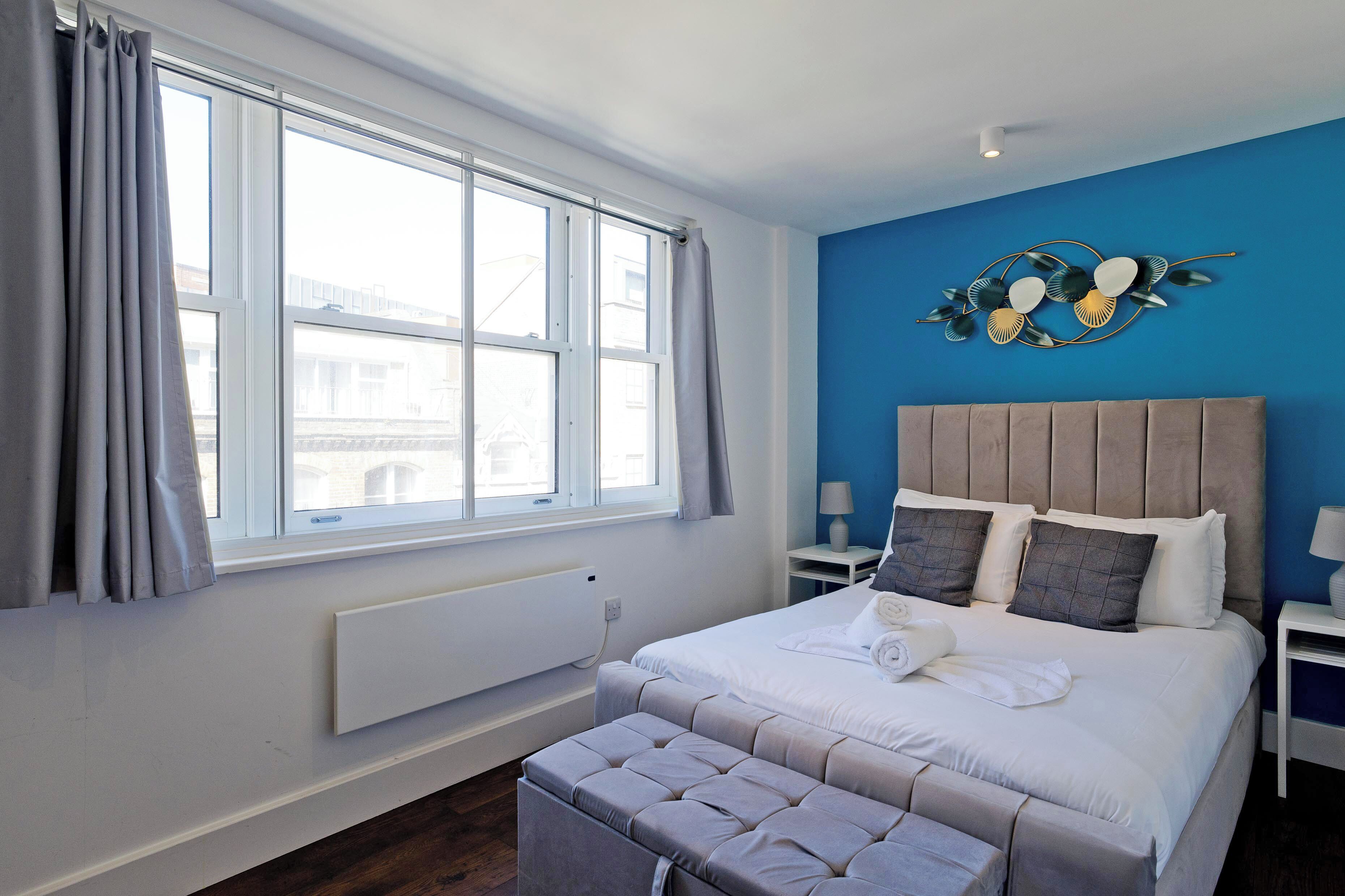A bedroom with a large window, a bed with a headboard, a bedside table, and a wall-mounted decoration.