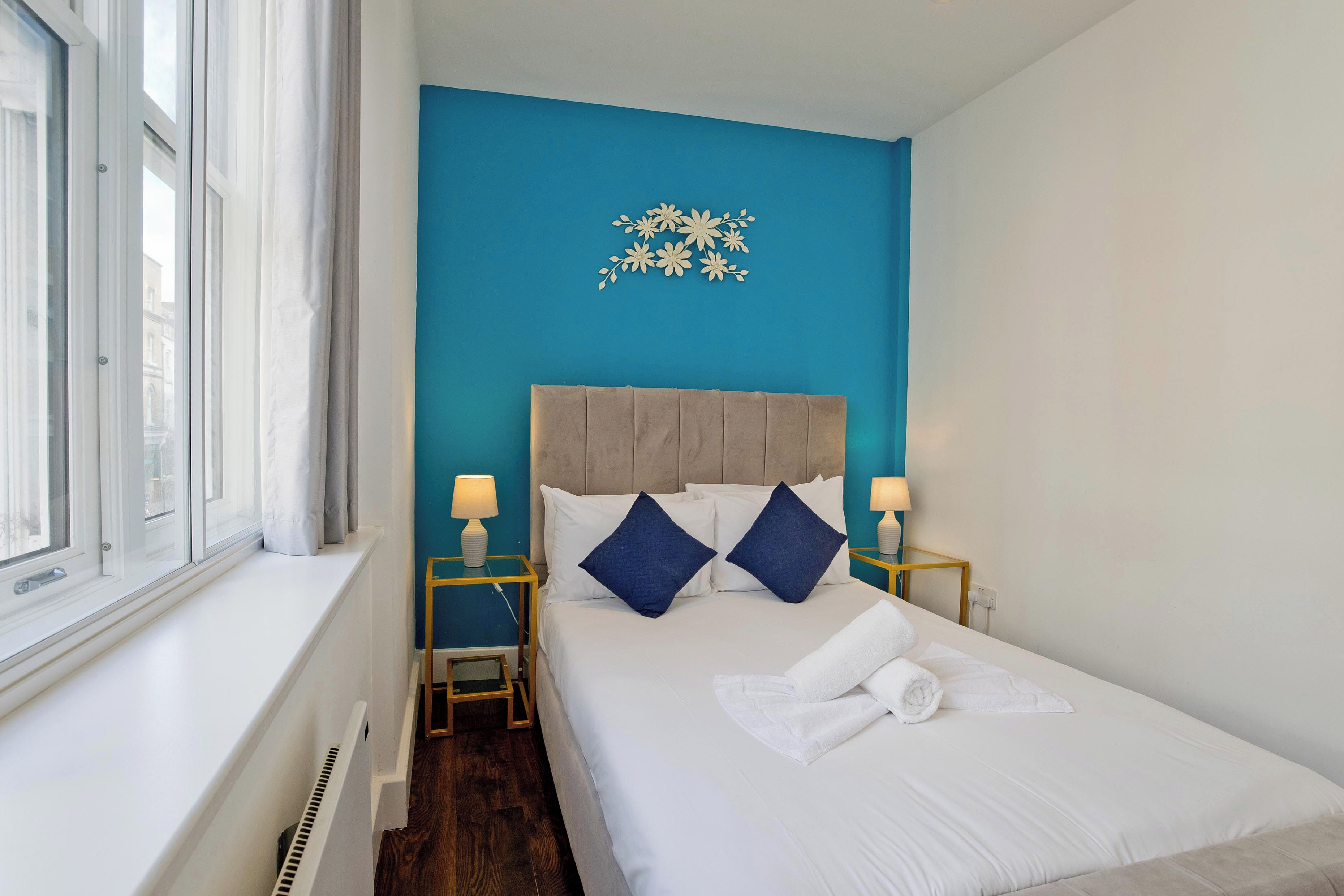 A hotel room with a bed, two bedside tables with lamps, a window with curtains, and a blue accent wall.