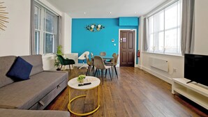 A modern living room with a sofa, a round coffee table, a dining area with chairs, a television, and large windows. - Imperial Middlesex Street Apartments (London)