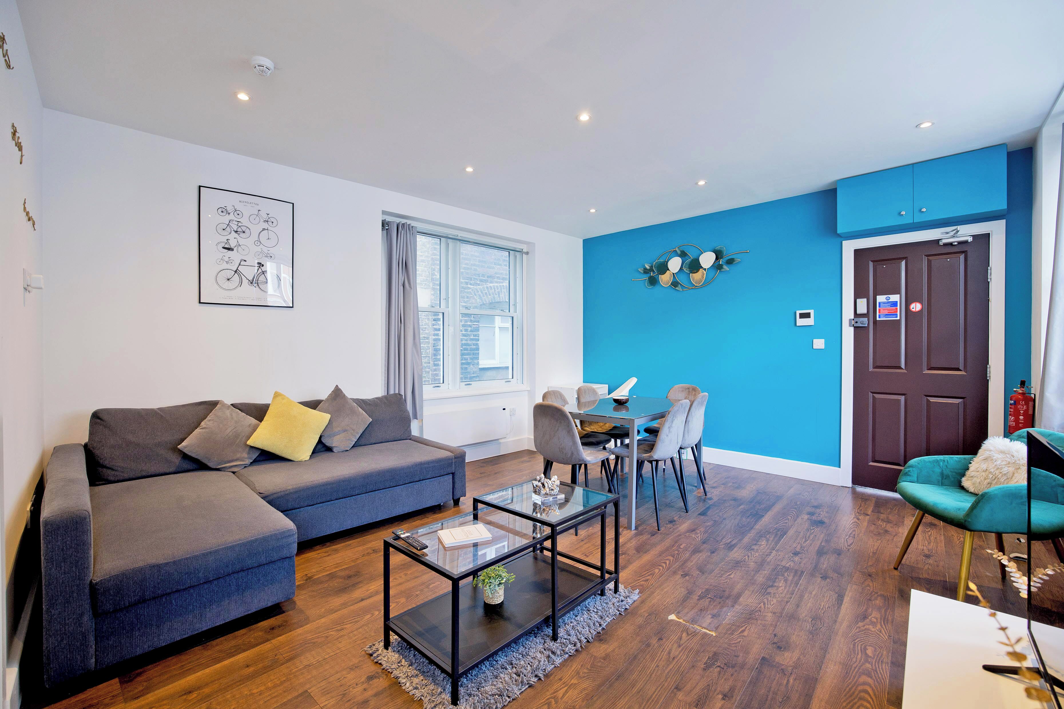 A modern living room with a sofa, coffee table, and chairs. There is a dining area with a table and chairs. The room has a blue accent wall and a door with a sign.