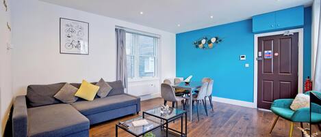 A modern living room with a sofa, coffee table, and chairs. There is a dining area with a table and chairs. The room has a blue accent wall and a door with a sign.