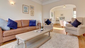 A living room with a beige sofa, two armchairs, a coffee table, and a rug. There are framed pictures on the wall and a kitchen area in the background. - Fabulous Seaside Retreat (Hunstanton)
