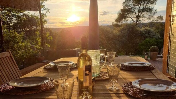 A wooden deck with a table set for dining, overlooking a scenic landscape with trees and a sunset.