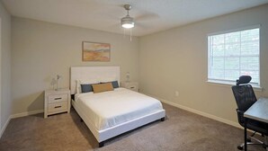 A bedroom with a bed, a desk, a chair, a ceiling fan, and a window.