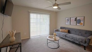 A living room with a grey sofa, a coffee table, a ceiling fan, and a TV mounted on the wall.