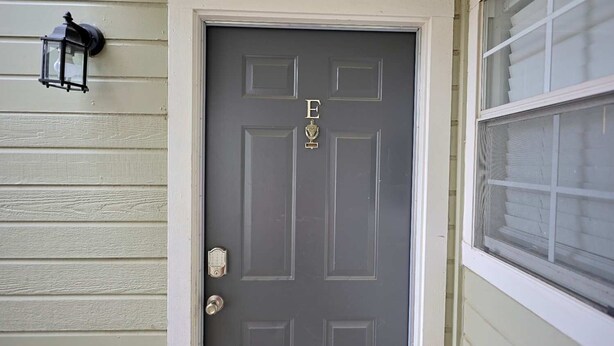 A gray door with the letters "E" and "D" and a trophy symbol, a black lantern on the wall, and a window with blinds.