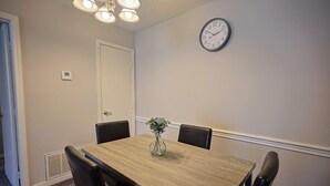A dining room with a wooden table, four chairs, a clock on the wall, and a door.
