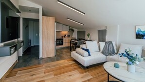 Una sala de estar moderna con un sofá, una mesa de centro y un televisor. Se observa una cocina al fondo con un comedor.