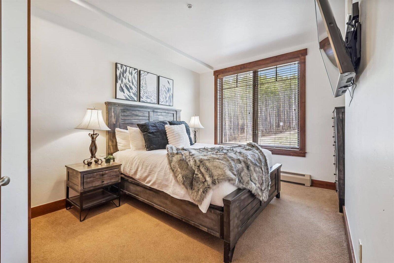 Une chambre à coucher comprenant un lit, des tables de chevet, des lampes, une grande fenêtre avec des stores et des œuvres d’art accrochées au mur.