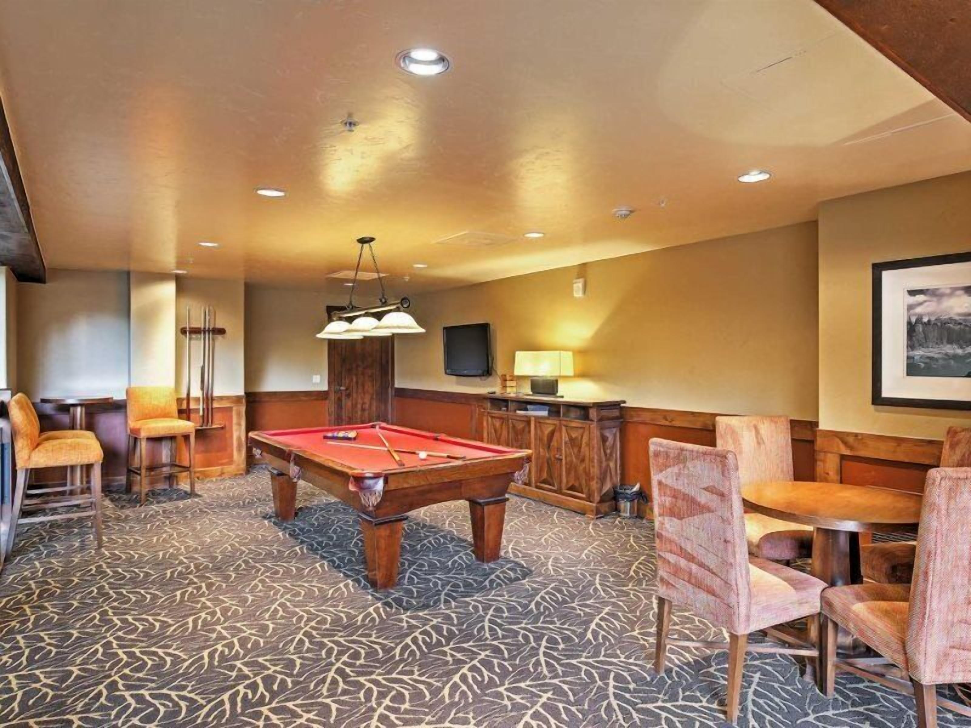 A pool table in a room with patterned carpet, wooden furniture, and a television on the wall.