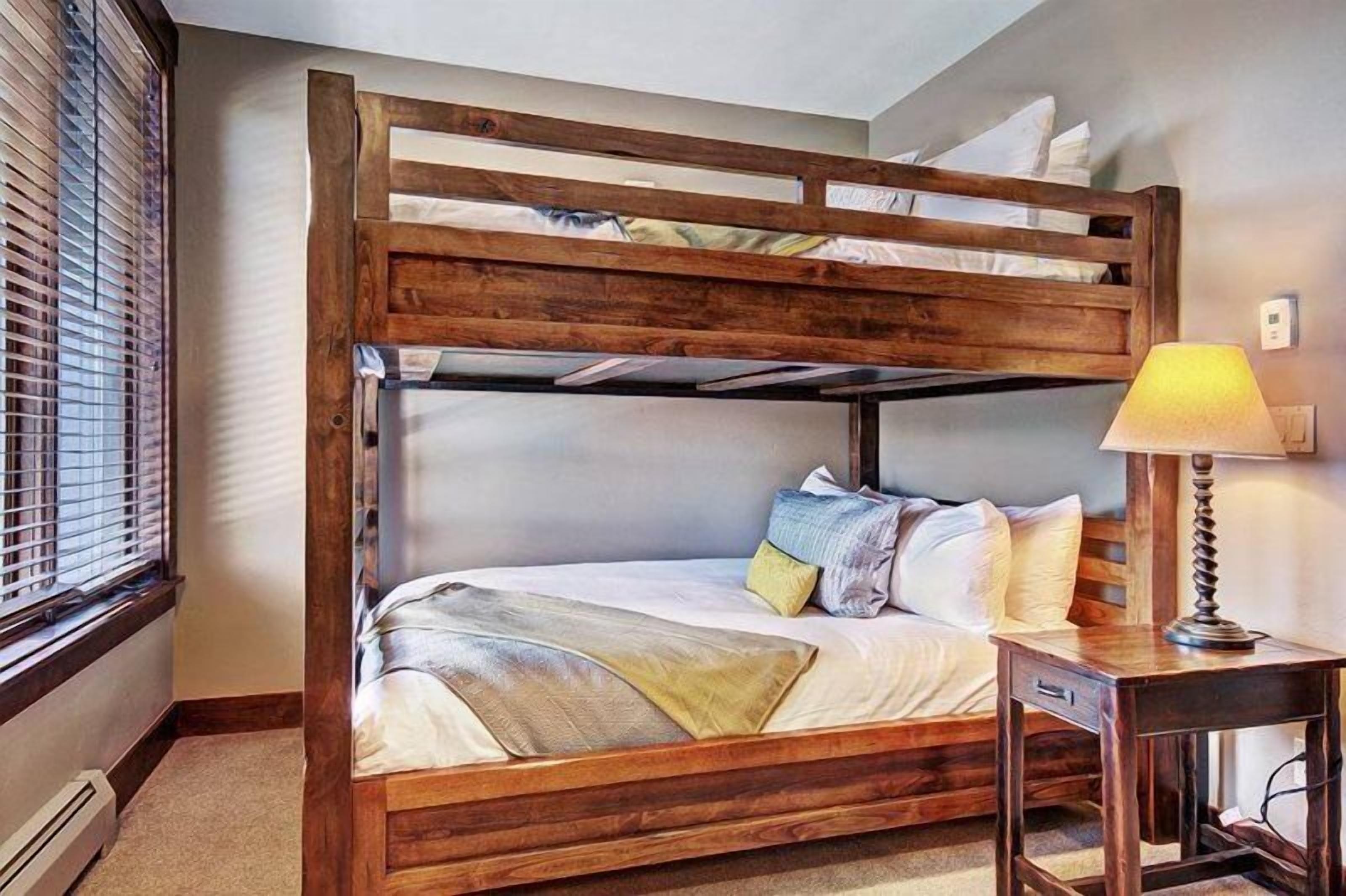 Un lit superposé avec une structure en bois, une table de chevet avec une lampe et une fenêtre avec des stores.