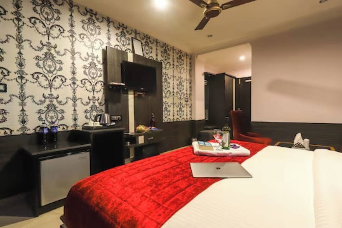 A hotel room with a red bedspread, a desk with a laptop, a TV mounted on the wall, and a ceiling fan.