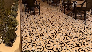 An outdoor patio area with tiled flooring, multiple tables and chairs, and wall-mounted lighting. - SelkA Pansiyon (Ayvalik)