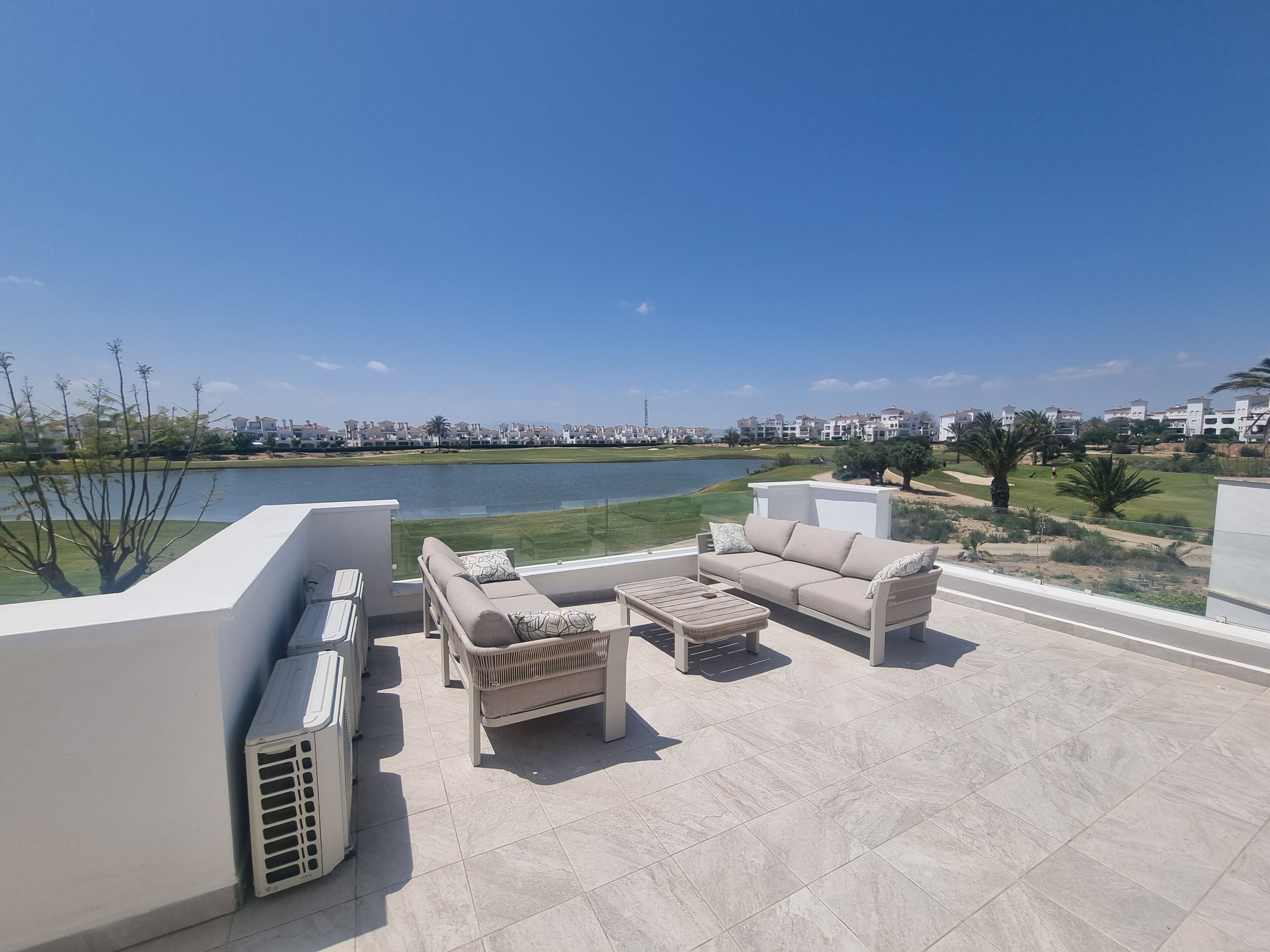 Una terraza en la azotea con mobiliario exterior, vista a un lago y una zona residencial al fondo.