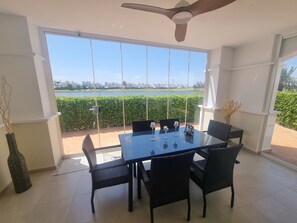 Uma área de refeições com mesa e cadeiras de vidro, janelas amplas com vista para um lago e edifícios, e um ventilador de teto.