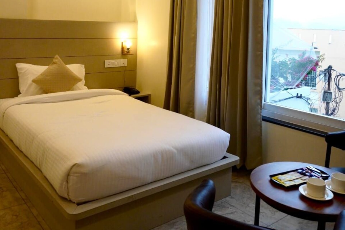 A hotel room with a large bed, a bedside table with a lamp, a small table with cups and a tray, and a window with curtains.