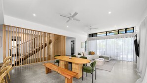 A modern living room with a wooden dining table, green chairs, a sofa, and a staircase with a wooden railing. - Villa Hitam - Burleigh Heads (Gold Coast)