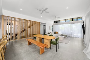 A modern living room with a wooden dining table, green chairs, a sofa, and a staircase with a wooden railing.
