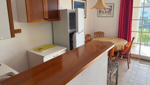 Cozinha compacta com armários de madeira, geladeira, mesa de jantar com cadeiras e janela com cortinas vermelhas.