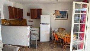 Uma cozinha compacta com armários de madeira, geladeira branca, mesa de jantar com cadeiras e vista para o exterior através de uma porta de vidro deslizante.