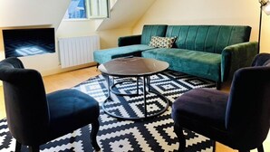 A cozy loft-style living room with a green sofa, two black armchairs, a coffee table, and a patterned rug. A television is mounted on the wall, and a floor lamp is visible. - La Colombage, Charming stay on the Place du Château (Amboise)