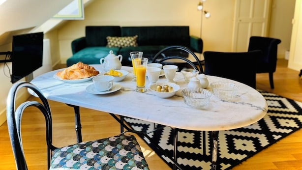 A dining table set with breakfast items, a patterned chair, and a black and white rug in a room with a sloped ceiling and a green sofa. - La Colombage, Charming stay on the Place du Château (Amboise)