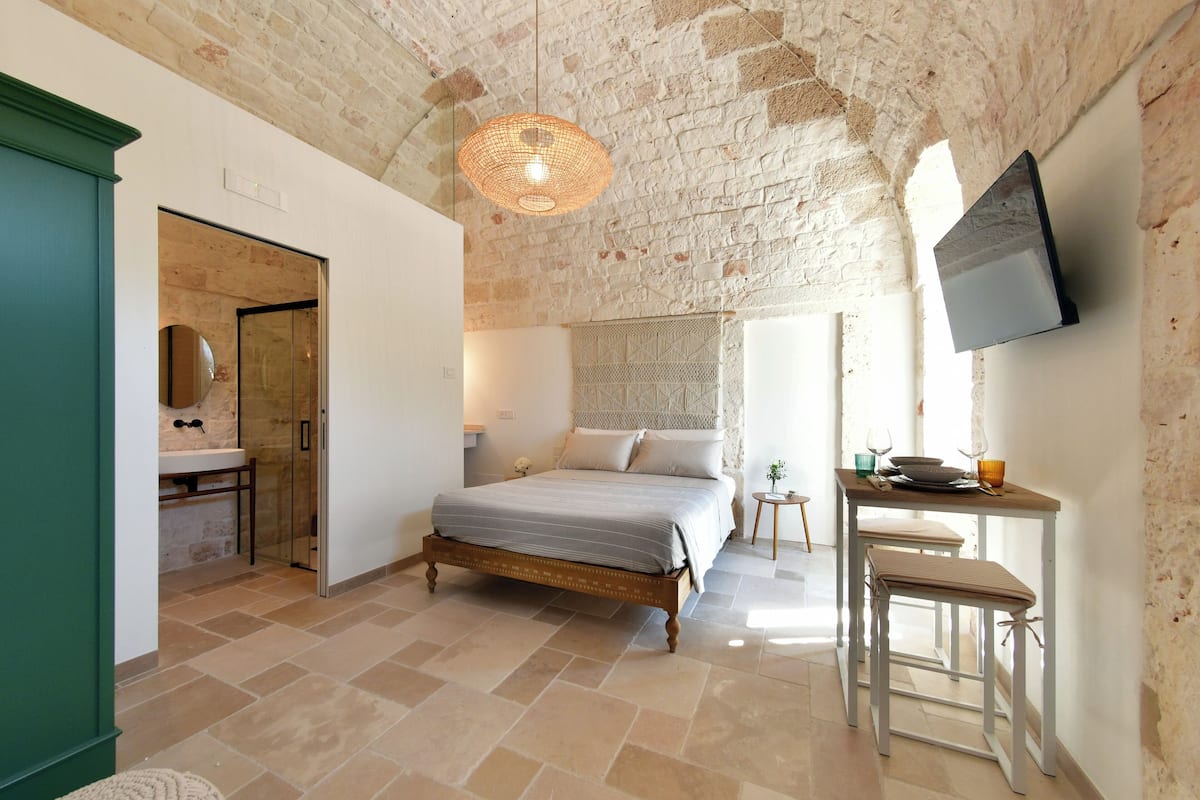 Quarto com parede de pedra, cama, mesa de cabeceira, televisão e janela.