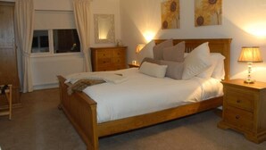 Un dormitorio con una cama de madera, dos mesitas de noche con lámparas, una mesita auxiliar, un ventanal con cortinas y dos cuadros colgados en la pared.