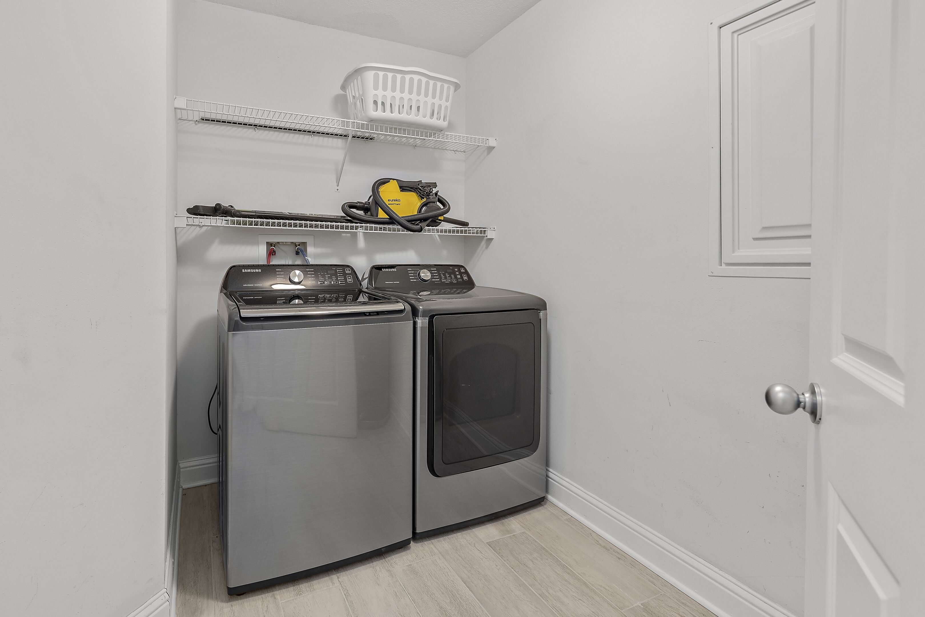 A compact laundry area with a washing machine and dryer, a white basket on a shelf, and a door to the right.
