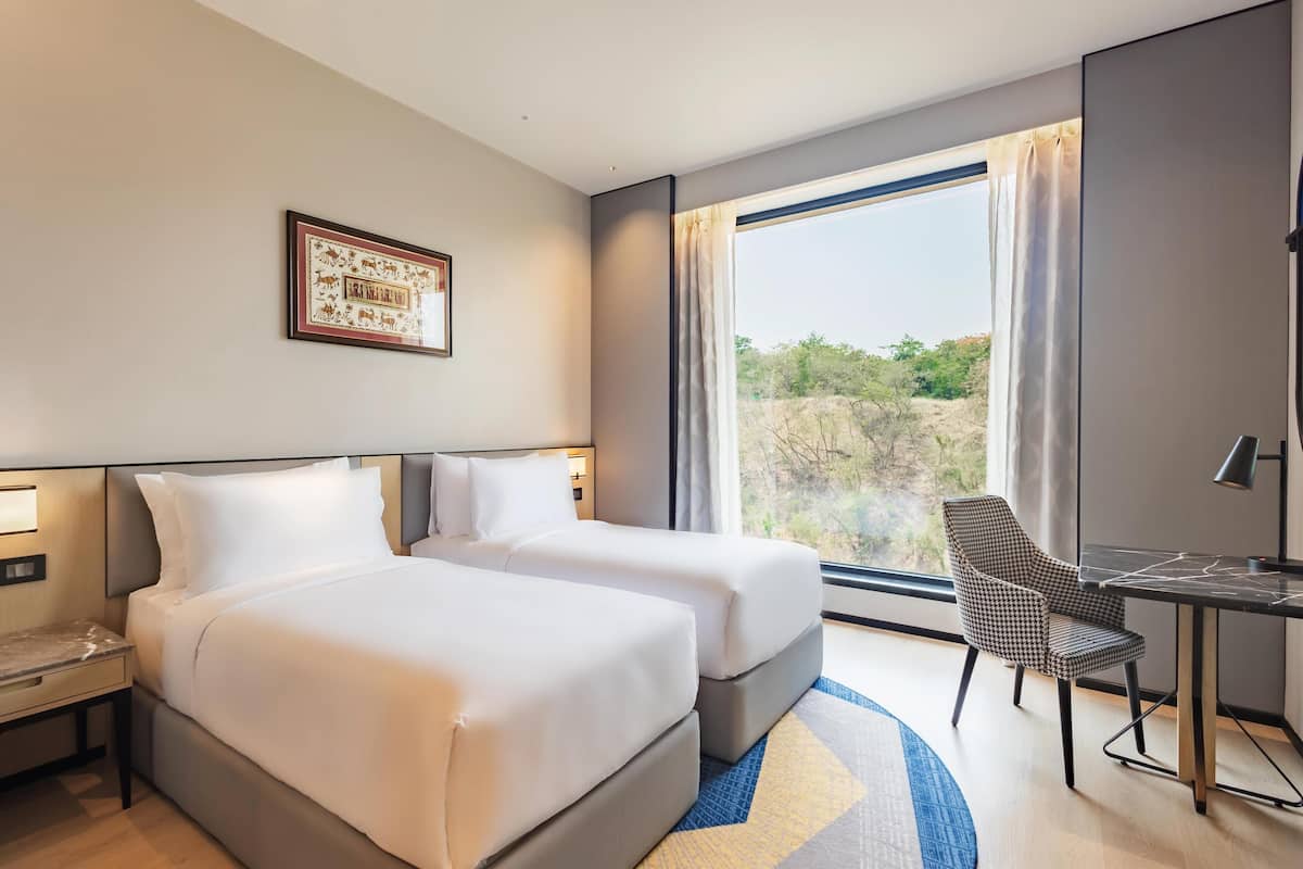 Habitación de hotel con dos camas, un escritorio, una silla, un ventanal grande con cortinas y un cuadro en la pared.