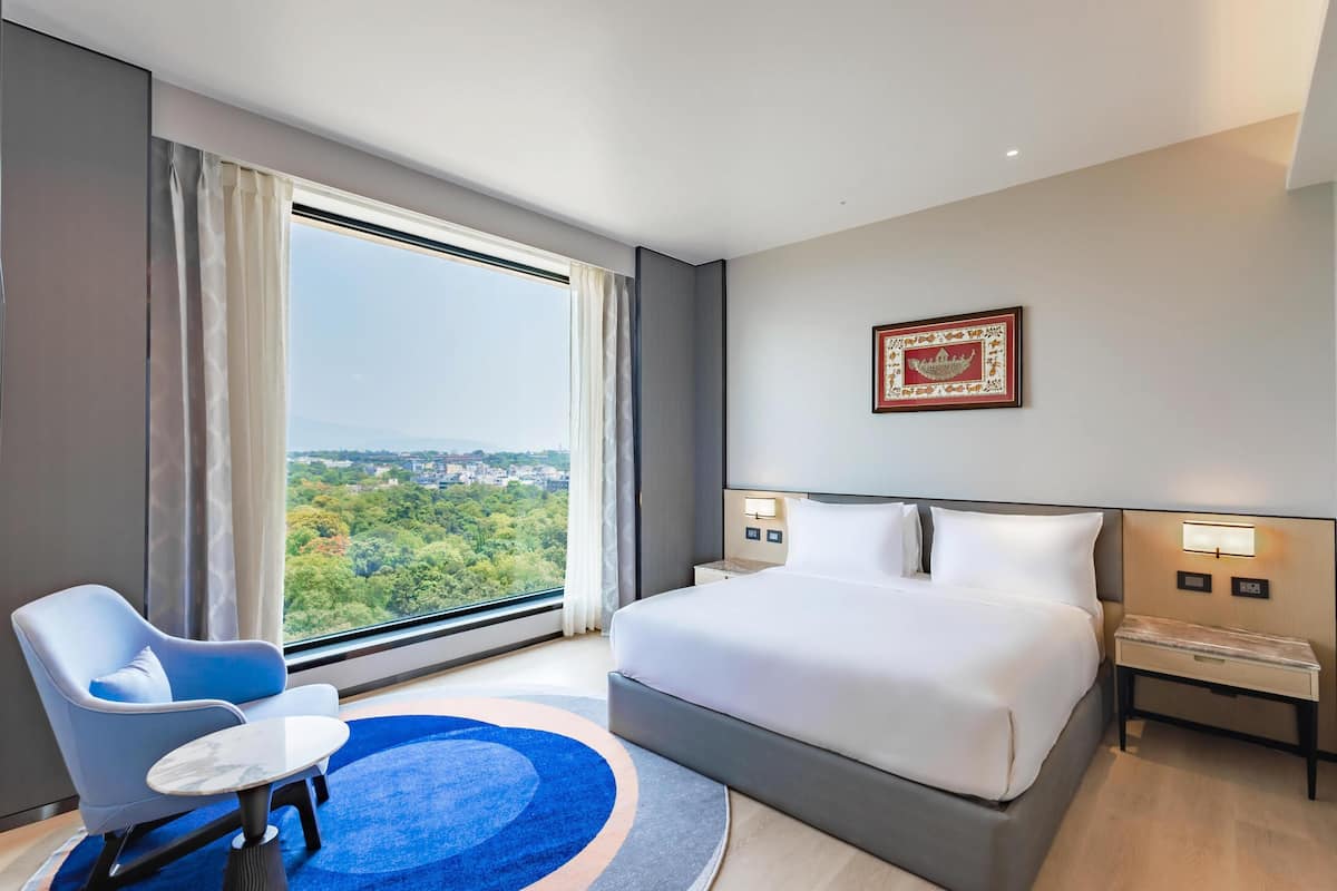 Una habitación de hotel moderna con una cama grande, una silla azul, una mesita pequeña y vistas de un paisaje urbano a través de un ventanal amplio.