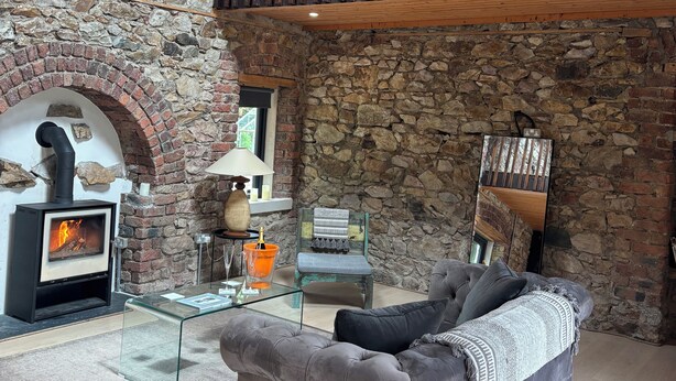 Un salon chaleureux avec un mur en pierre, une cheminée, une table basse en verre et un canapé gris.