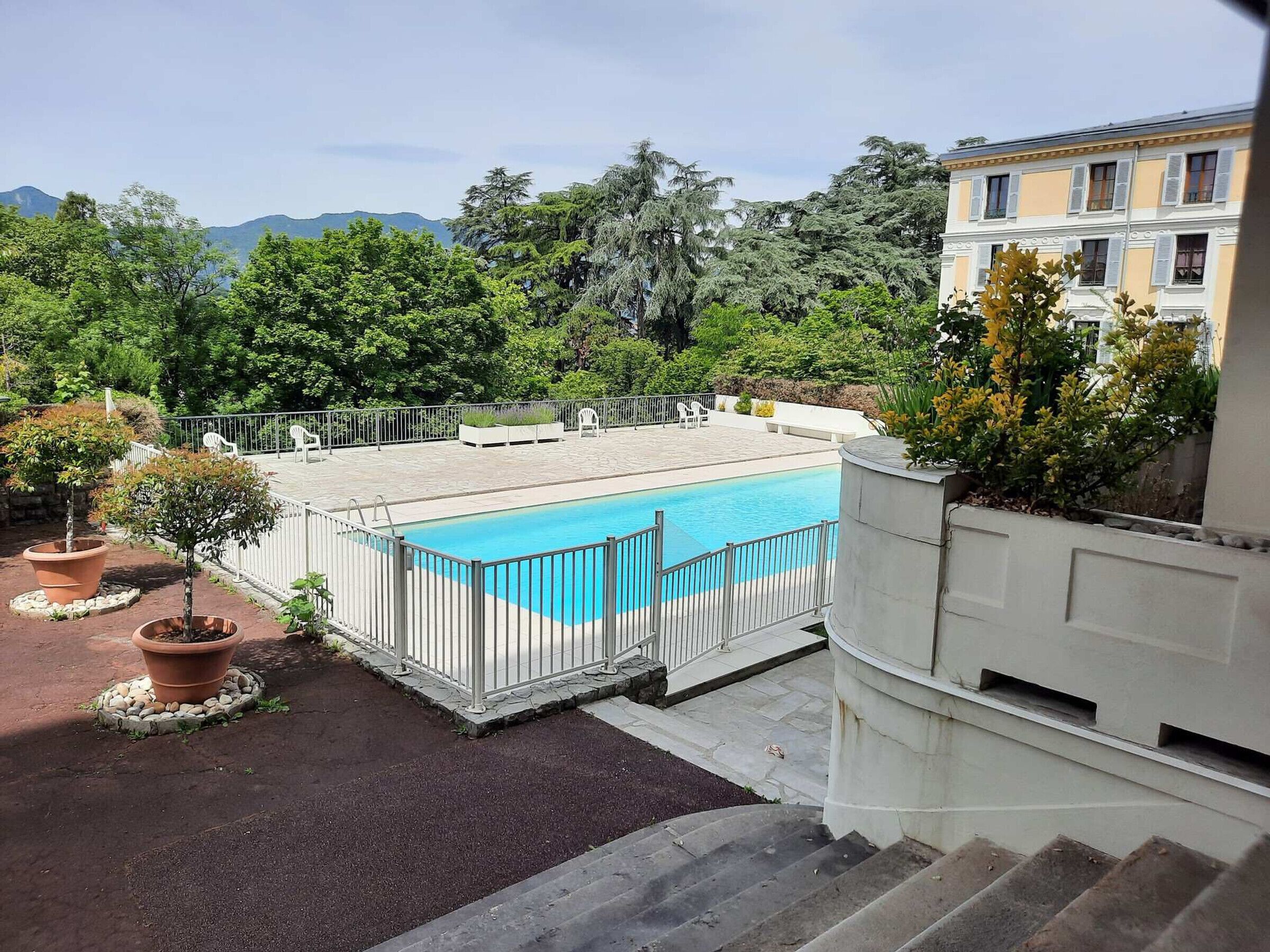 A rooftop pool with a white fence, surrounded by potted plants and a view of a building with a balcony.