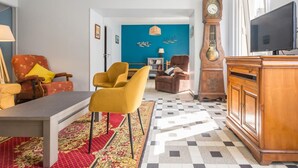 Uma sala de estar com piso xadrez, um armário de madeira, uma televisão, um relógio e duas cadeiras.