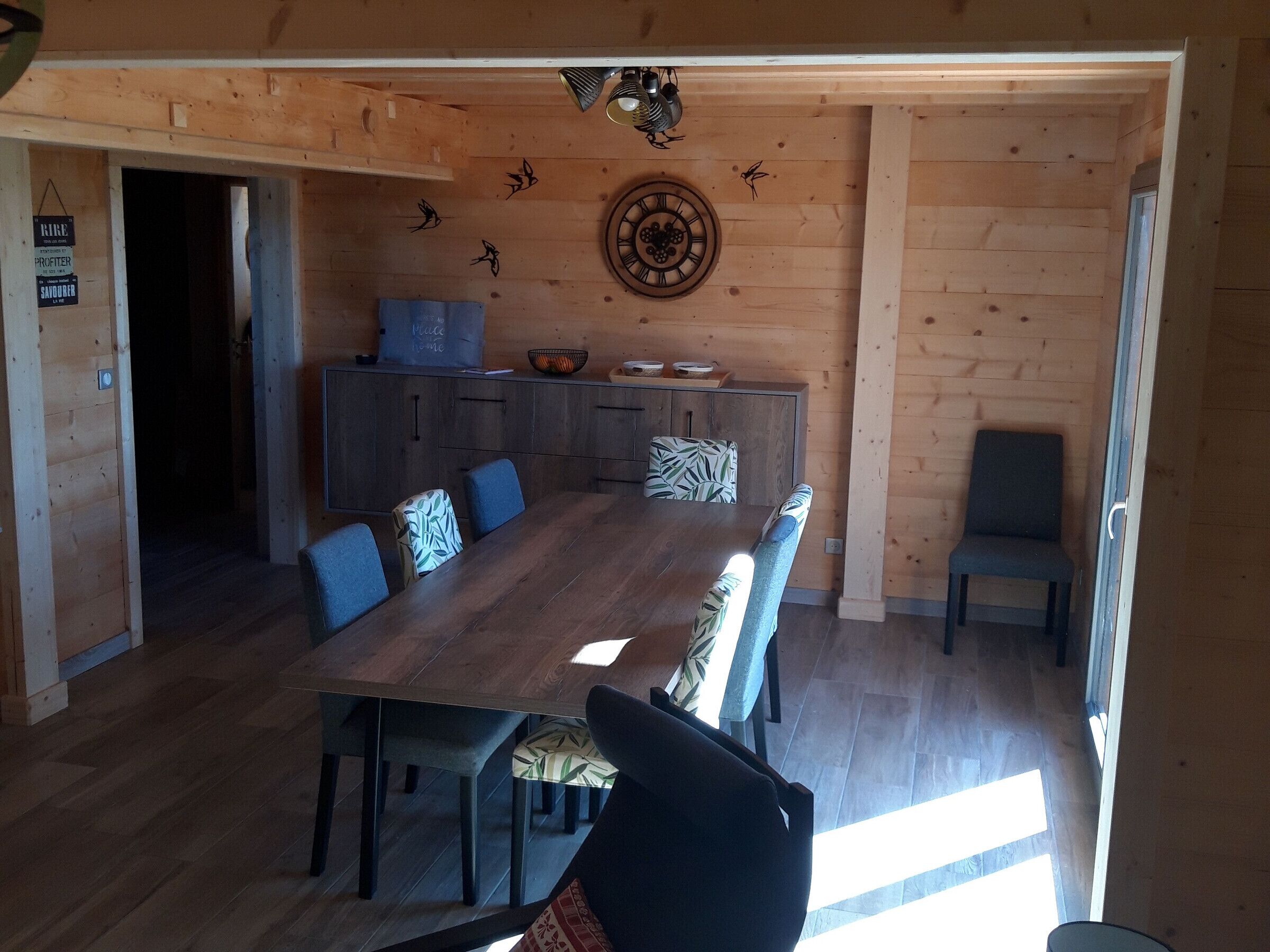 A wooden interior with a dining table, chairs, a kitchen area, and a decorative wall clock.