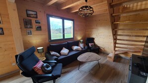 Een gezellig chaletinterieur met een houten vloer, een bank, twee stoelen, een kleine tafel en een trap.