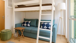 A bunk bed with a ladder, a teal sofa with patterned pillows, a small wooden table, a green ottoman, and a floor lamp. - StayINN - Leipzig, BSS25a (Leipzig)