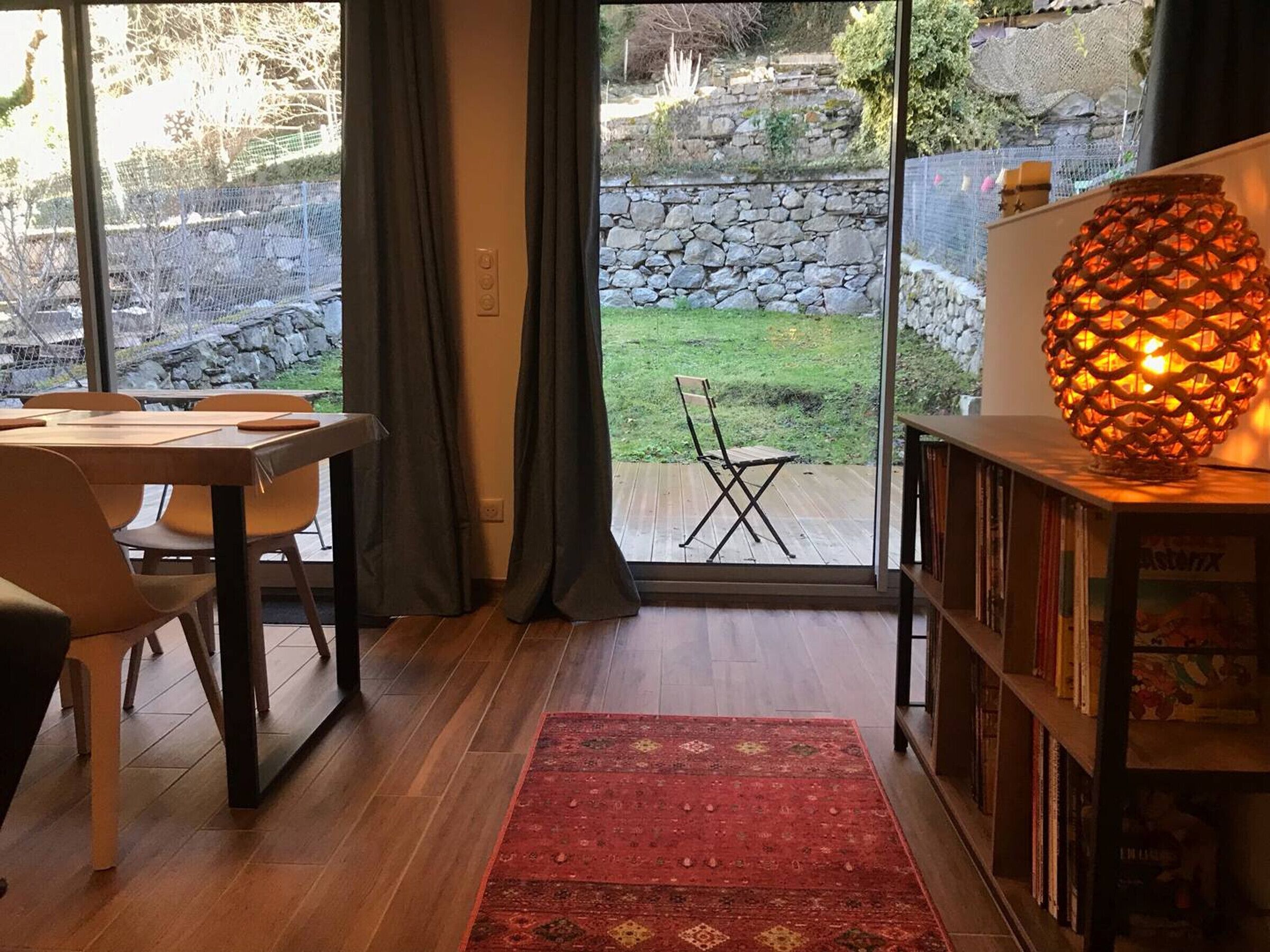A modern living room with a wooden floor, a red rug, a dining area with a table and chairs, a bookshelf, and a large window with curtains.