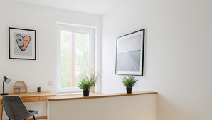 Un escalier avec des rampes en bois menant à un palier où se trouvent un bureau et une chaise, une fenêtre ornée de plantes et des œuvres d’art encadrées accrochées aux murs.