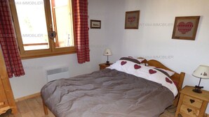 Un dormitorio con una cama, una mesita de noche, una ventana con cortinas, un calefactor de pared y un cuadro enmarcado.