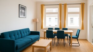 A modern living room with a blue sofa, a wooden coffee table, and a dining area with chairs and a table. - Apartment Halle city center - 3 rooms (Halle (Saale))