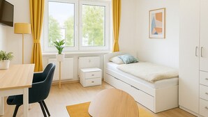 Et lille værelse med seng, natbord, stol, skrivebord og garderobeskab.