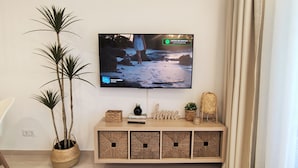 Un salon avec un téléviseur à écran plat fixé au mur, un fauteuil blanc, une console sur laquelle sont disposés des objets décoratifs, et deux plantes en pot.