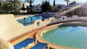 Une piscine divisée en deux zones distinctes, l’une avec un bord incurvé et l’autre avec un bord droit. On y trouve des chaises longues et un espace ombragé avec des parasols.
